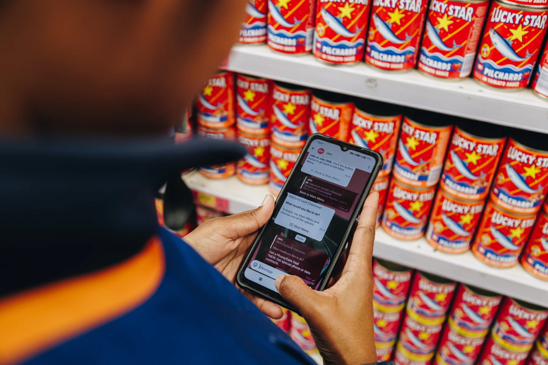 A supermarket employee in uniform standing at a shelf of tinned fish, looking at the Jem chatbot’s main menu on their phone.