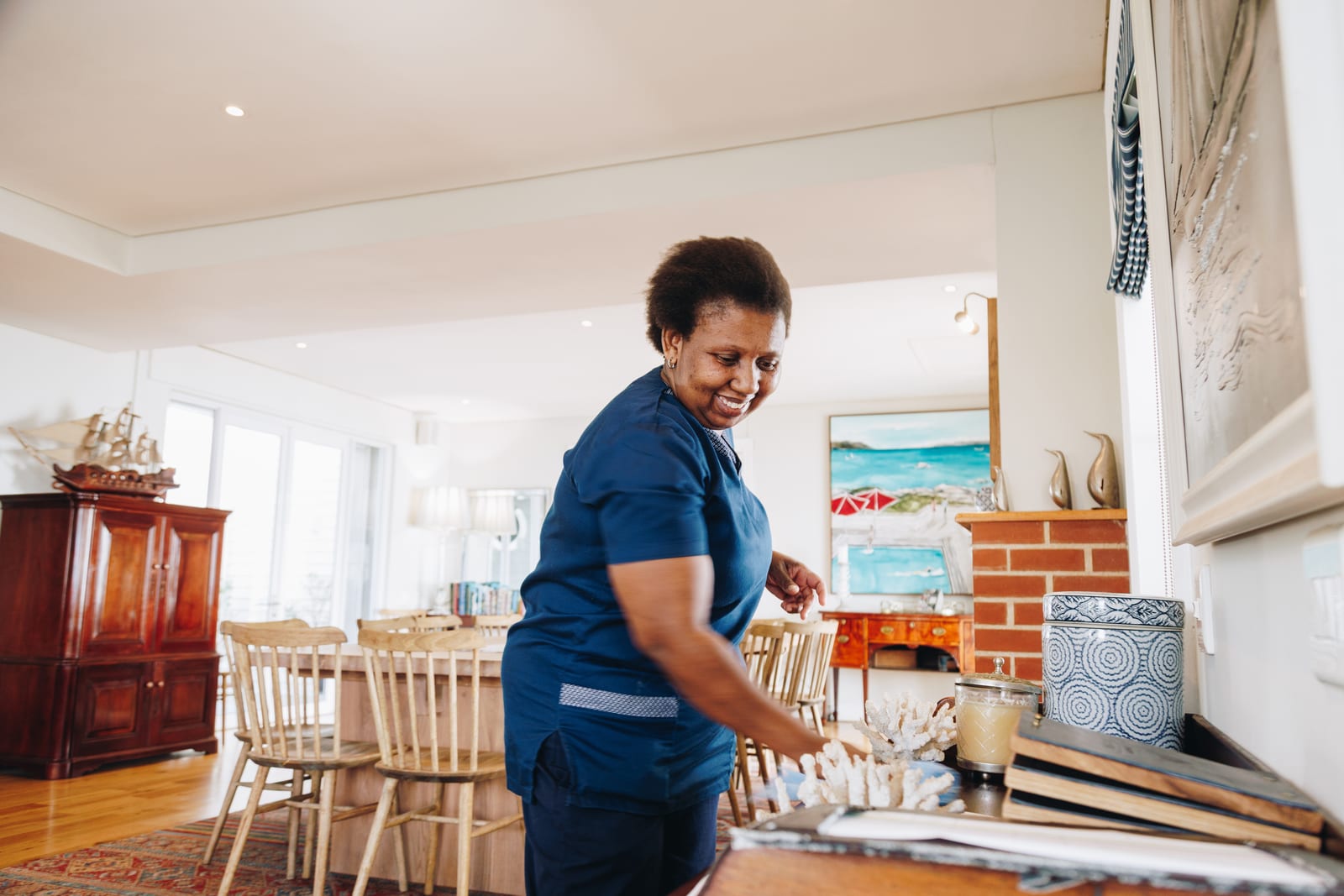 A housekeeper in a navy uniform smiling as she dusts a mantelpiece in a bright, furnished living room