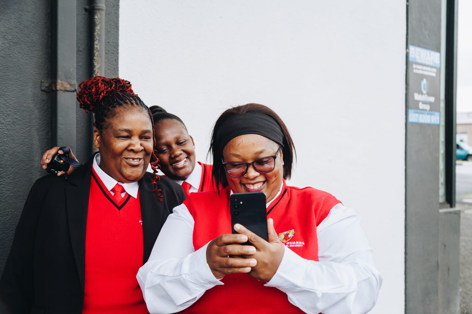 Three security guards in red uniforms crowded around one phone outside a building, all laughing at what is on the screen