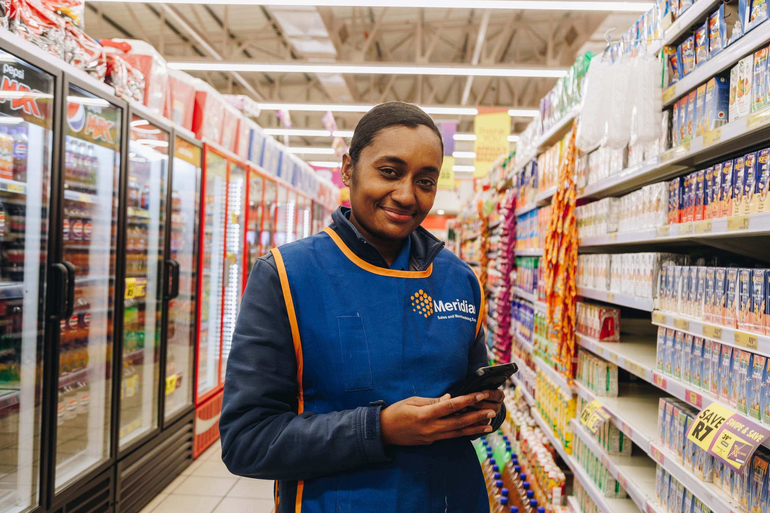 A Meridian Group merchandiser facing up product on a supermarket aisle, phone in hand — one of the 5,500 deskless people this story is about
