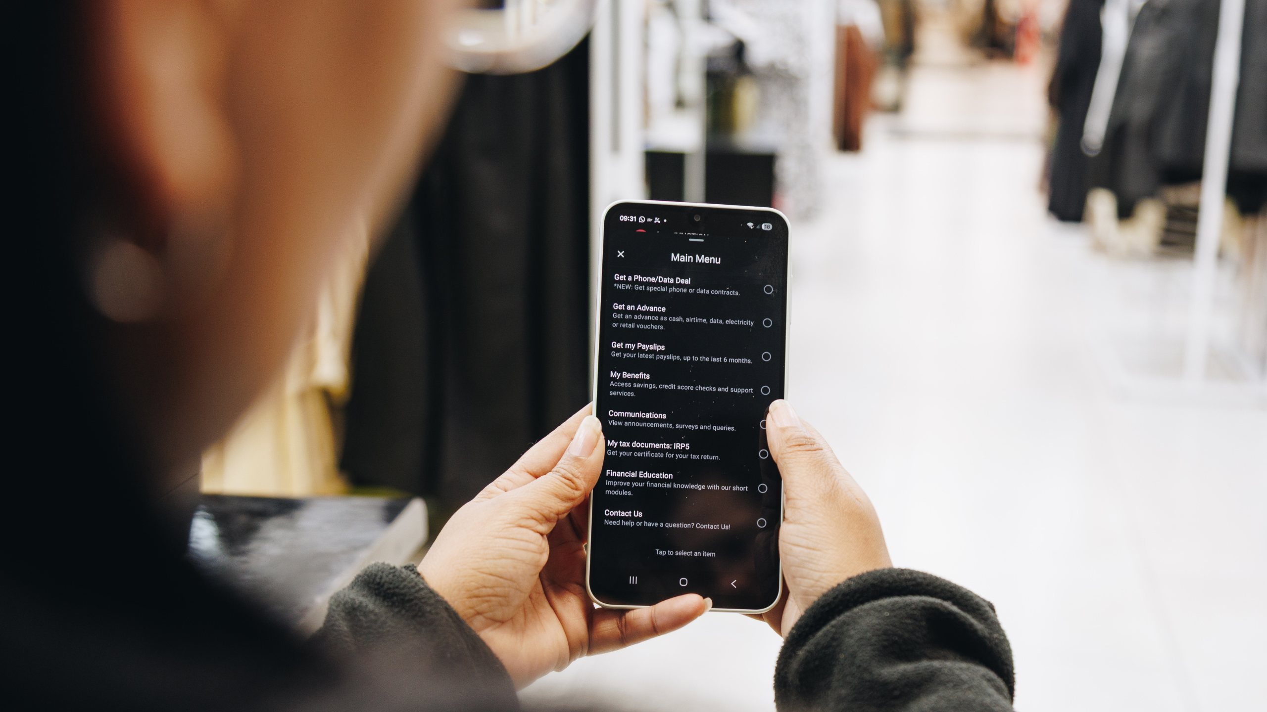 A shop worker holding a phone showing the Jem main menu, clothing rails stretching away behind her