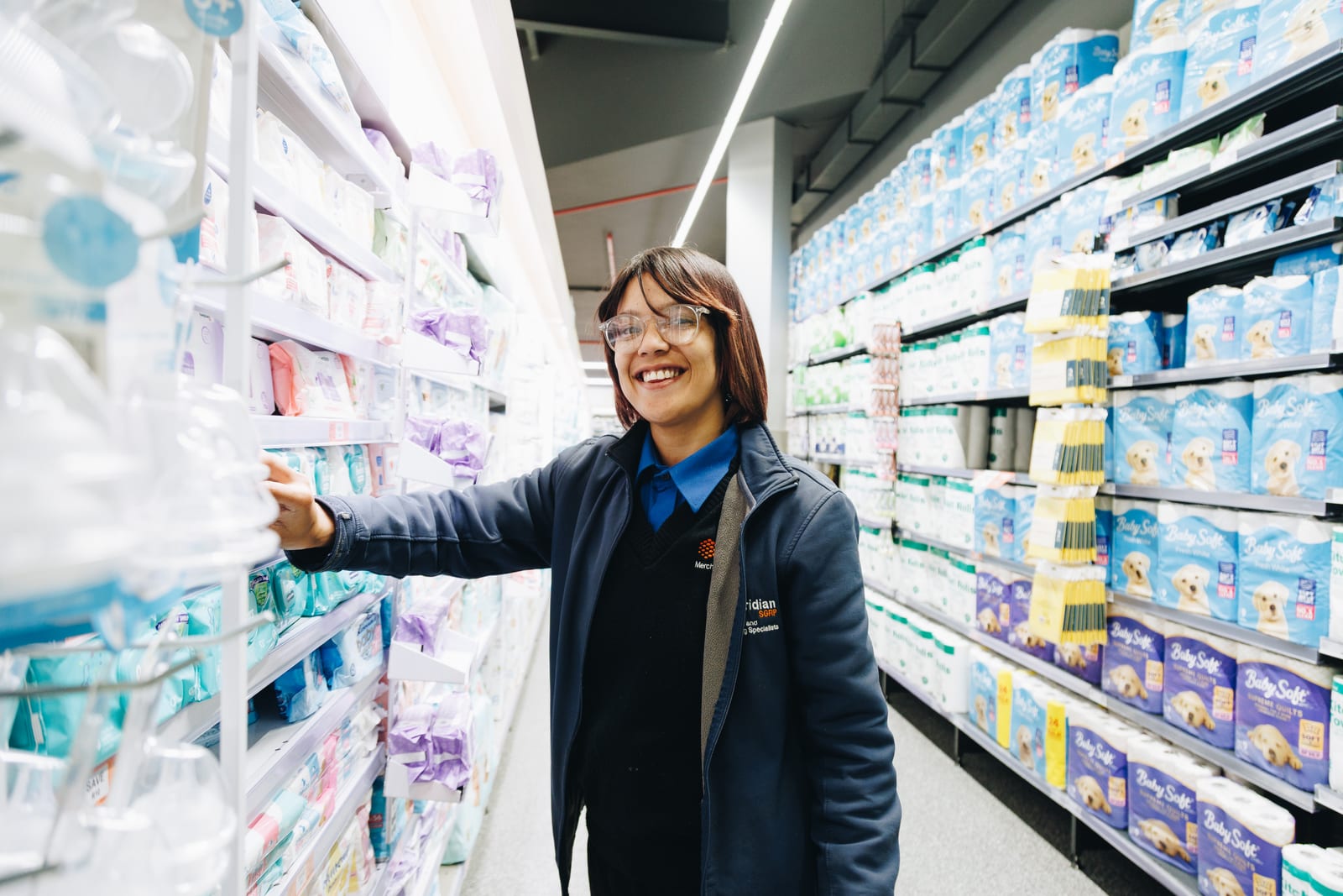 A Meridian merchandiser in a branded jacket smiling as she reaches for stock in a supermarket aisle