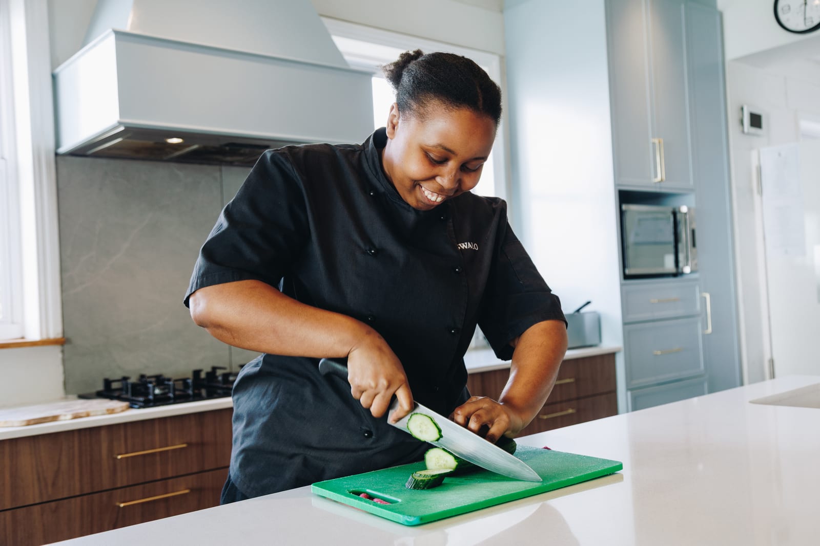 A chef in black kitchen whites smiling as she slices a cucumber on a green board in a bright kitchen