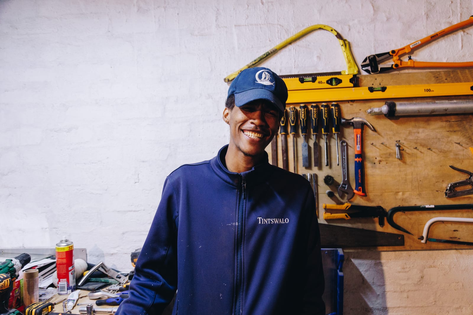 A maintenance worker in a navy Tintswalo jacket and cap smiling in a workshop, hand tools hanging on the board behind him